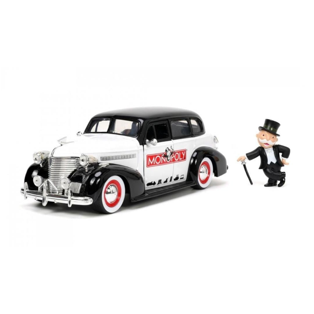 A diecast model of a classic 1939 black and white Monopoly car with red and white wheels, shown next to a miniature Sir Richard De Mille figurine on a white background.