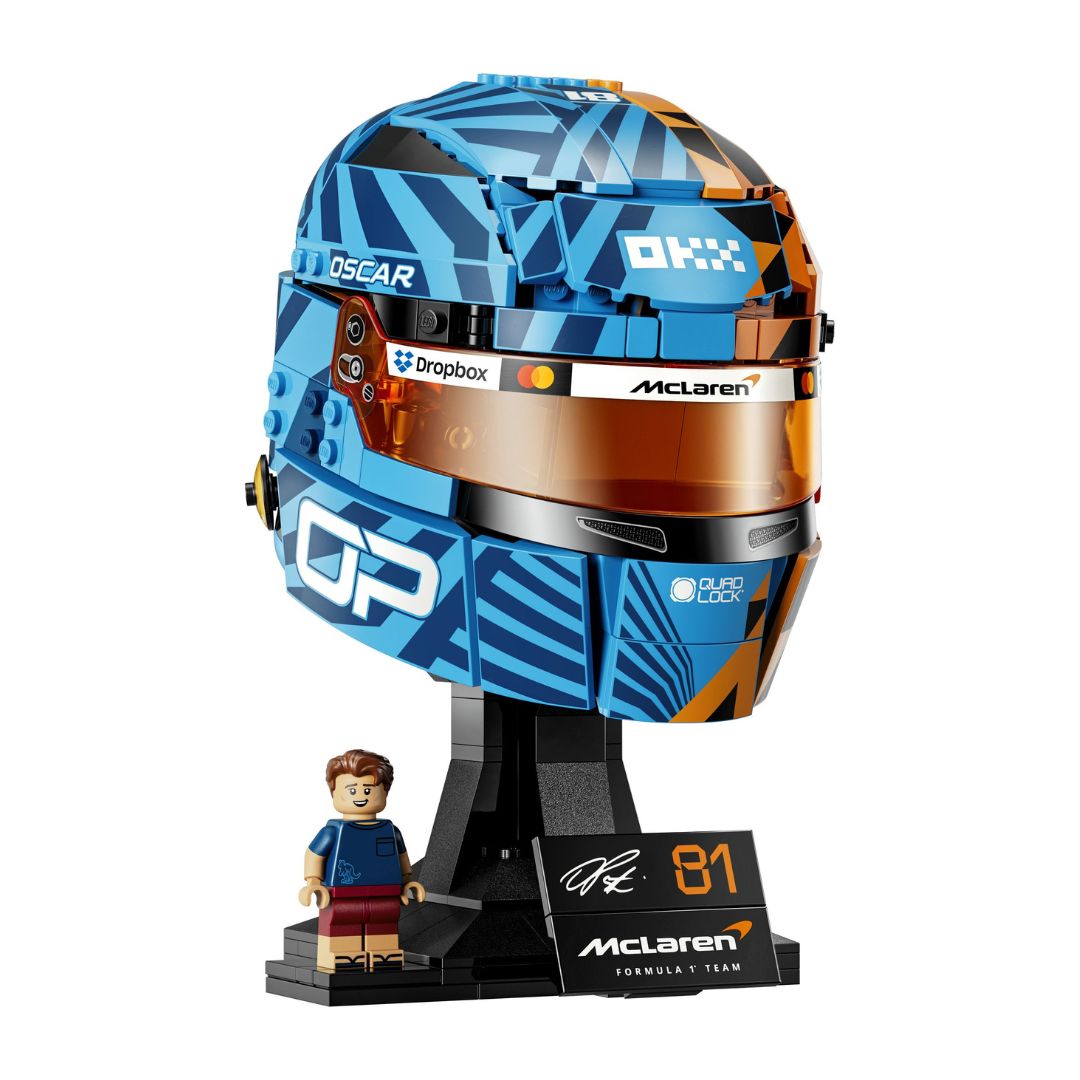 A building block model of a McLaren F1 Oscar Piastri helmet in blue, orange, and black, displayed on a black stand with a small minifigure and a plaque featuring the number 81 and McLaren Formula 1 Team branding.