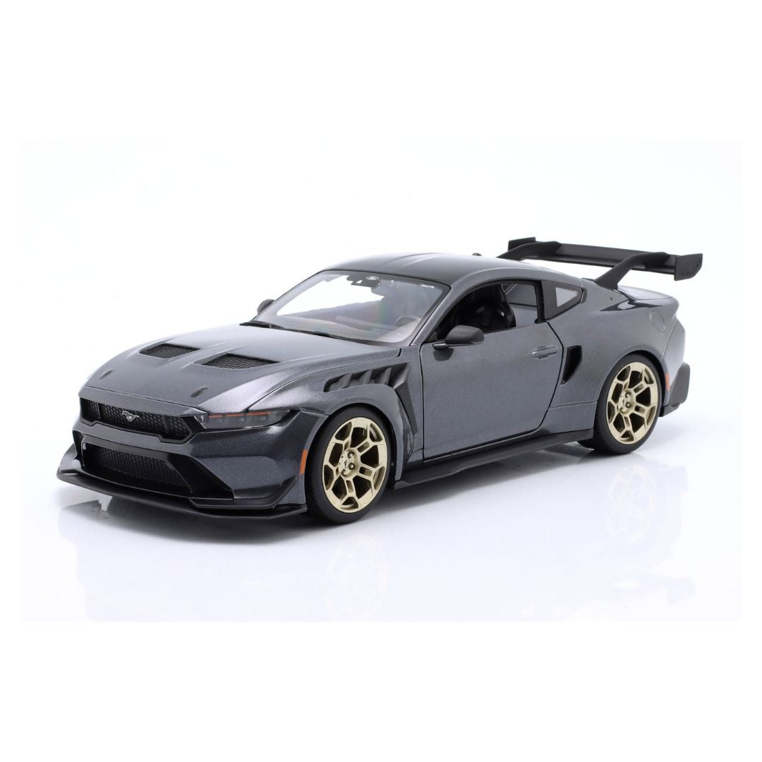 A dark gray die-cast model of a Ford Mustang with a widebody kit, gold multi-spoke wheels, and a rear spoiler, photographed on a white reflective studio background.
