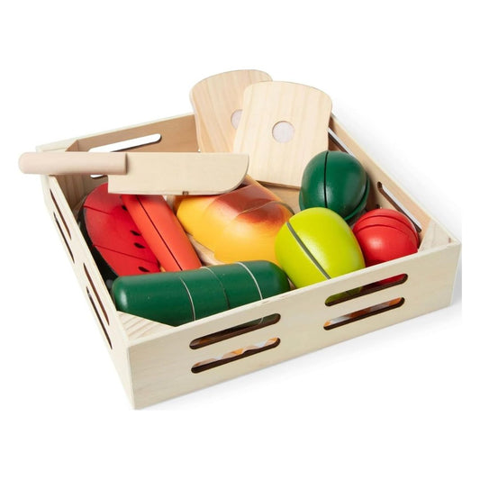 A wooden toy vegetable and fruit play set arranged inside a light natural wood crate on a white background. The set includes colorful plastic vegetables and fruits such as green and red radishes, a yellow-orange pear, and a lime, along with wooden cutting boards and a knife.