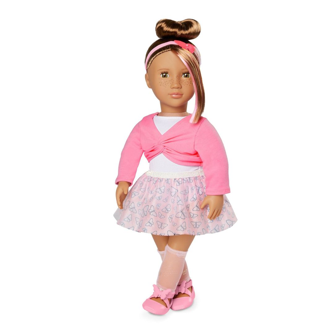 An 18-inch doll with brown skin and brown hair styled in a high bun, wearing a pink long-sleeve top, a light pink patterned skirt, pink socks, and pink ballet flats, set against a plain white background.